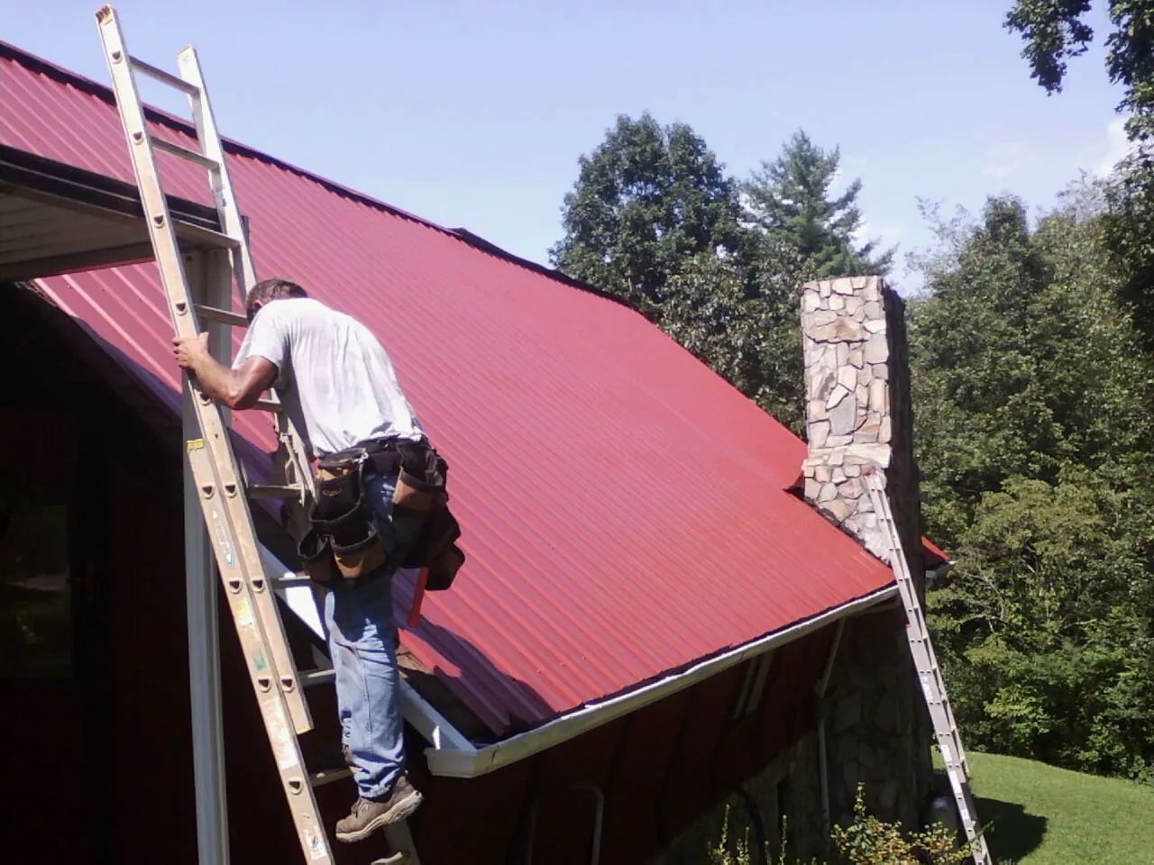 Roofer on a ladder beside a red metal roof and stone chimney — Soffit & Fascia Repair by Roofing Brothers in North Branford